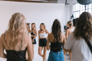 professional photo studio workshop, clean white backdrop, softbox lighting, group of newfaces practicing poses with a creative director, attendees watching and learning, makeup artist table in the background, healthy model lifestyle elements