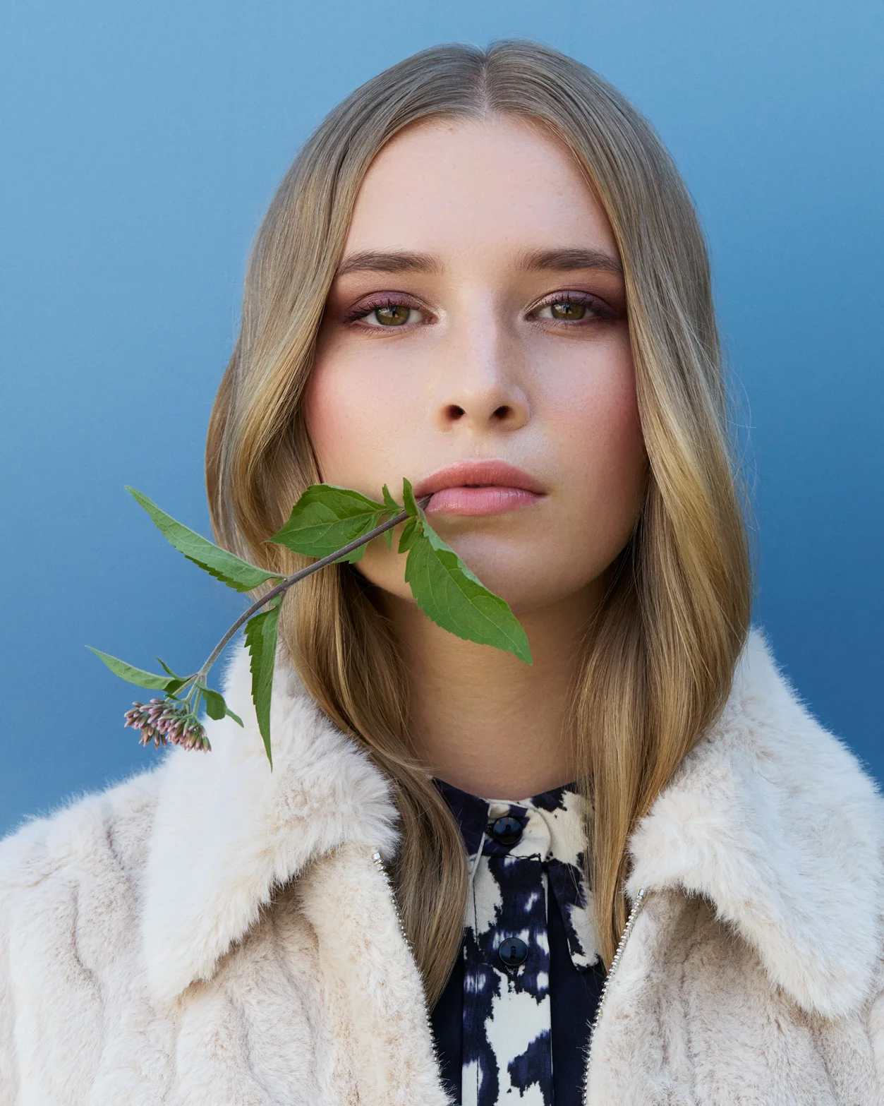 Close-up von Paula mit natürlichem Make-up und grünem Blatt im Gesicht, vor blauem Hintergrund.